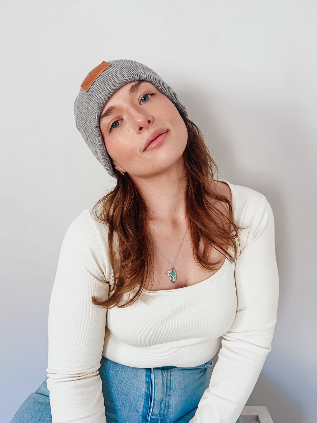 Woman wearing a gray beanie with brown leather patch, white long-sleeve top, and blue jeans against a plain background.