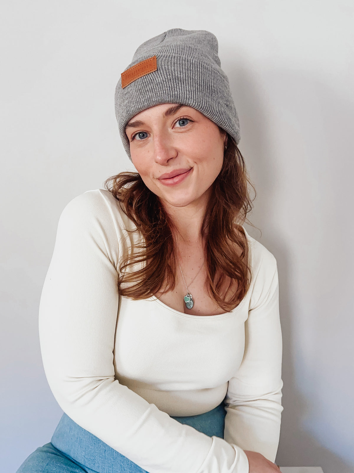 Woman wearing a gray knit beanie with a brown patch on a plain background